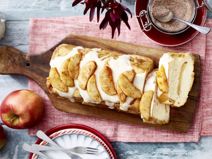 Auf einem Brett liegt ein Apfel-Biskuit-Kuchen aus der Kastenform. Er ist überzogen mit Vanillesoße und mit Äpfeln dekoriert. 