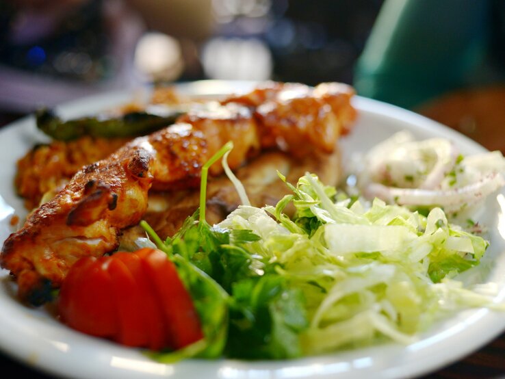 Türkische Hähnchenspieße Tavuk Şiş vom Grill mit Salat auf weißem Teller. Im Hintergrund verschwommene Tiscdekoration. Frontalansicht. 