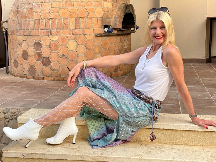 Renate Zott sitzt lächelnd auf einer steinernen Treppe und hat ein lebhaftes Outfit an. Sie hat blonde Haare, trägt ein weißes Tanktop und darüber eine bunte, schimmernde Hose mit einer Netzstrumpfhose darunter und weiße Stiefeletten.